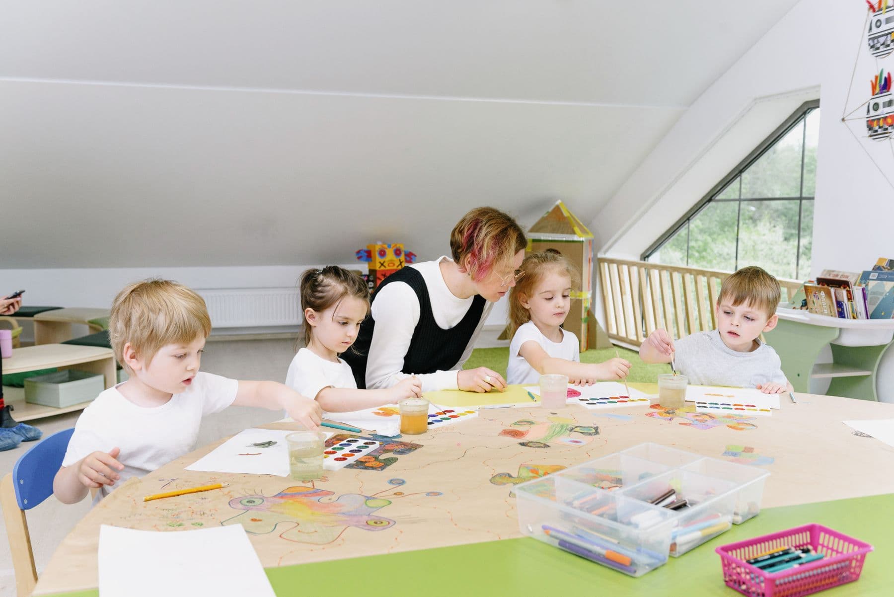Four young children painting and drawing at a shared table alongside their teacher