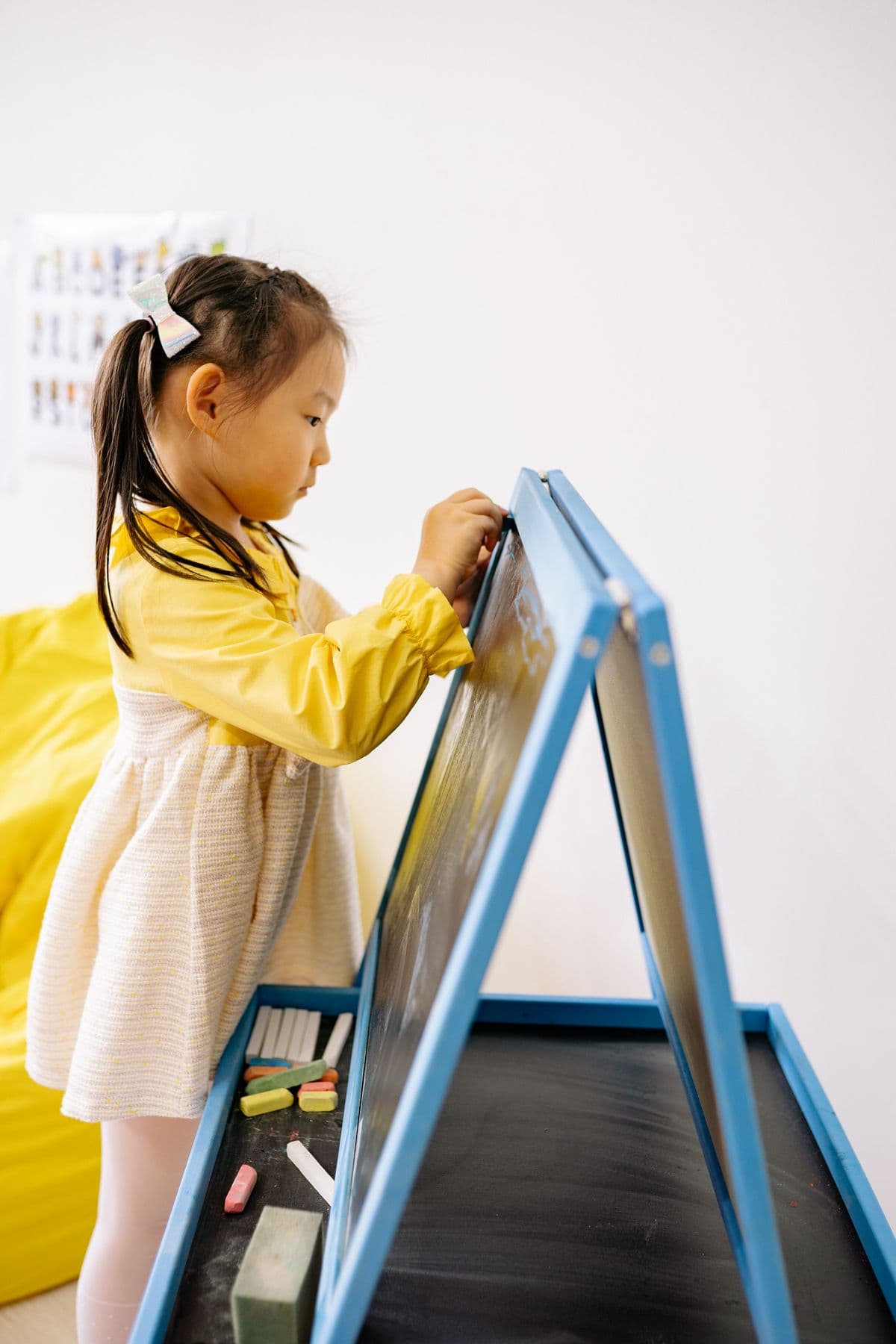 A young girl in a yellow dress writing in chalk on a small easel blackboard