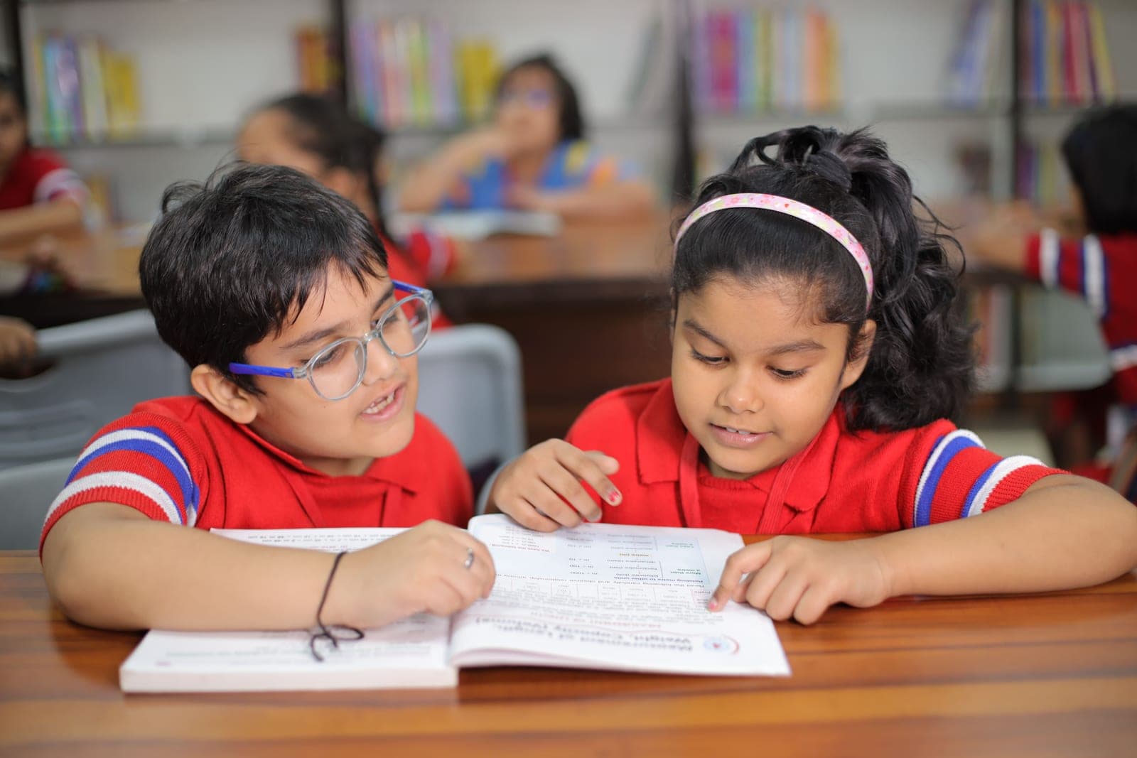 Two primary-age children in red school uniform reading a workbook together at a desk